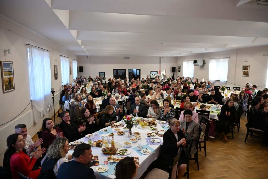 Widok na salę w Domu Ludowym w Jabłonce, przy zastawionych stolikach siedzą kobiety oraz mężczyźni zaangażowani w organizację uroczystości.